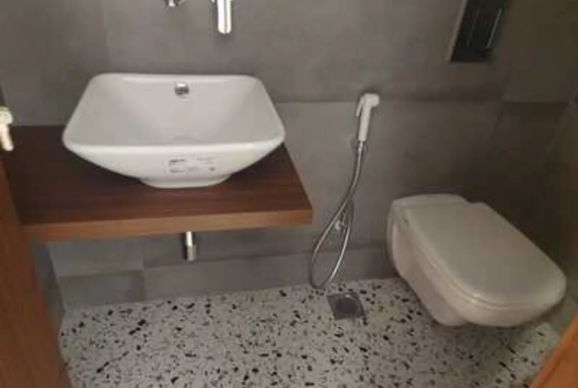 Modern bathroom with square washbasin, wooden shelf, wall-mounted toilet, and speckled flooring in a rental apartment in Greater Kailash-1, South Delhi.