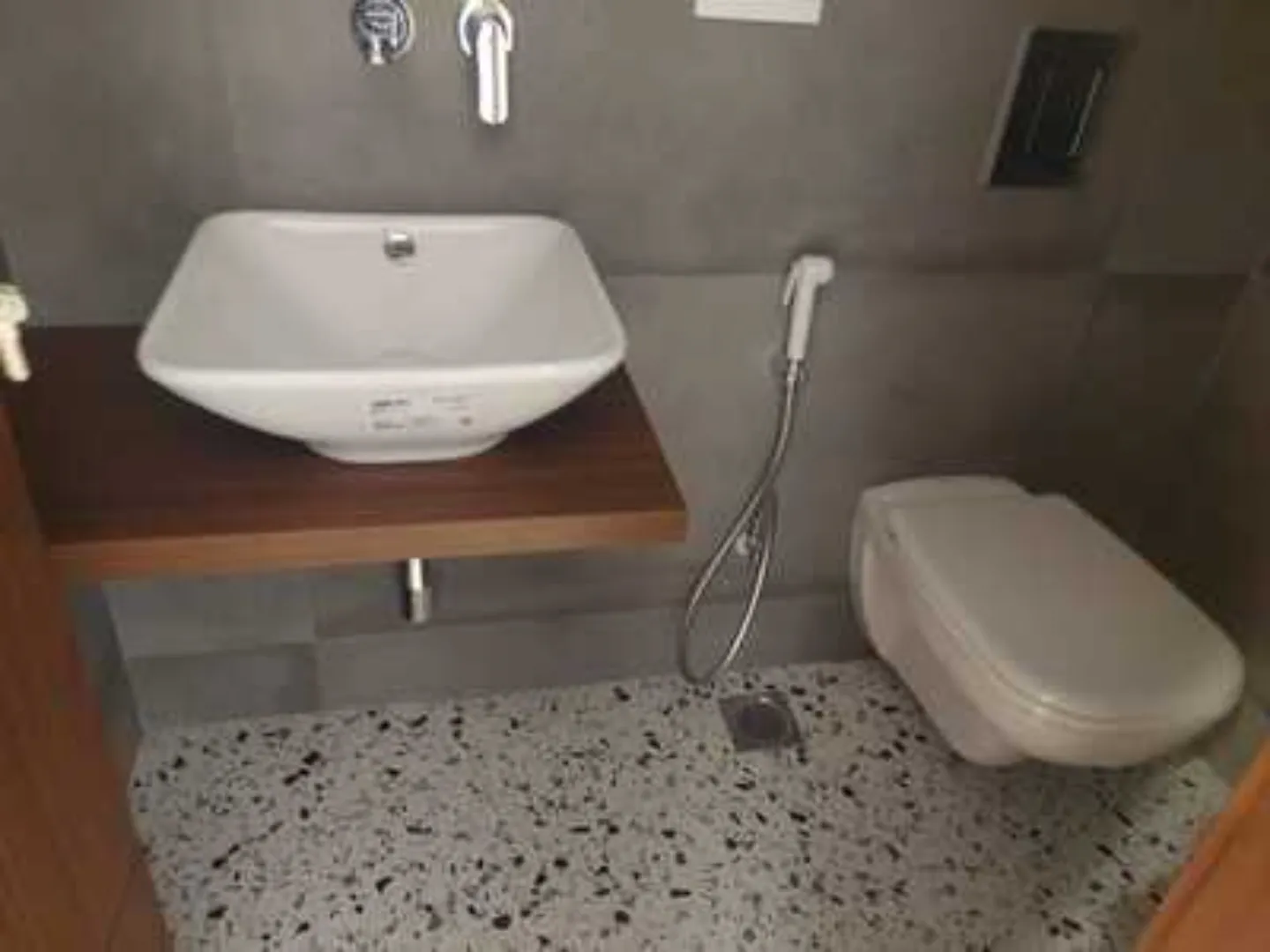 Modern bathroom with square washbasin, wooden shelf, wall-mounted toilet, and speckled flooring in a rental apartment in Greater Kailash-1, South Delhi.