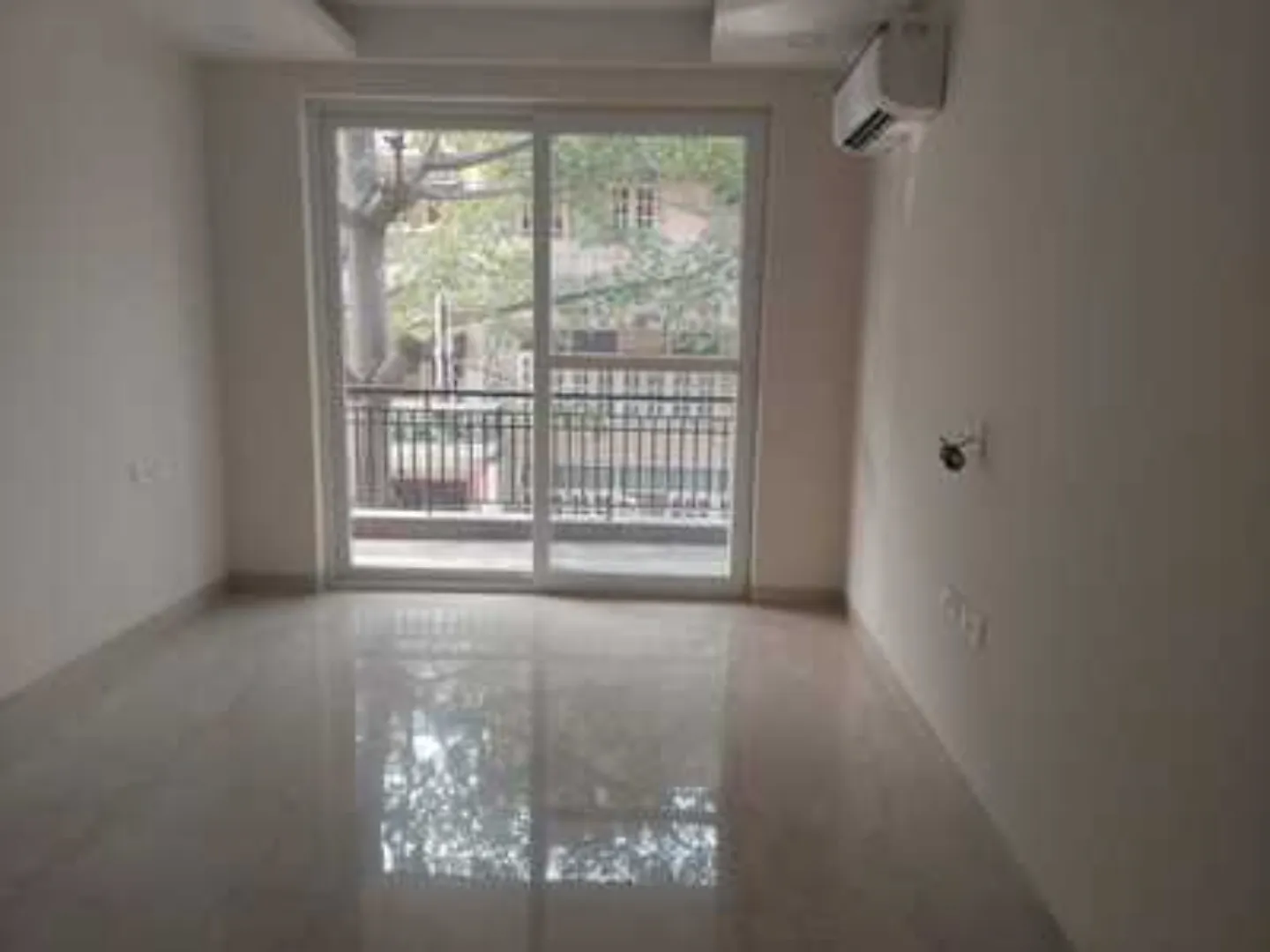 Empty room with sliding glass doors to a balcony, polished floor tiles, and AC unit in a rental apartment in Greater Kailash-1, South Delhi.